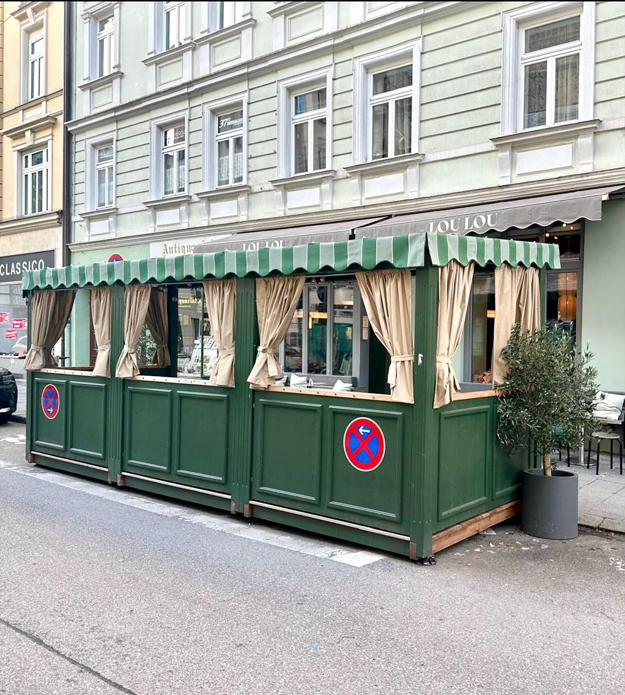 Fertiger Schanigarten des Lou Lou in München — dunkelgrüne Einfassung mit Streifenmarkise und Olivenbaum